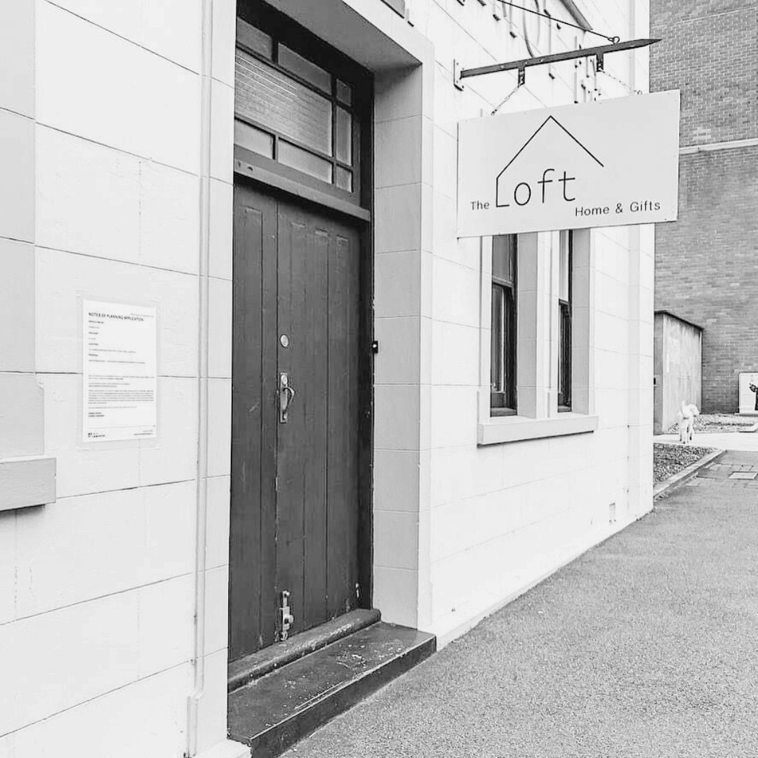 black and white street image of dark door and hanging sign that says the loft home and gifts