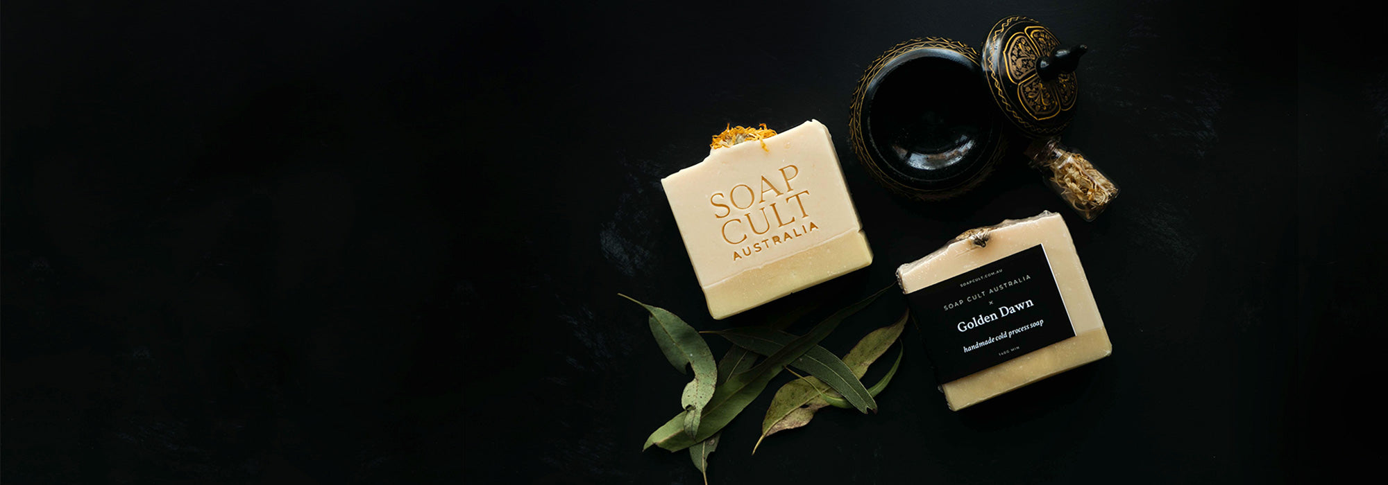 On a black background, two yellow and cream handmade soap bars in minimal occult aesthetic packaging sits with australian made gumleaves