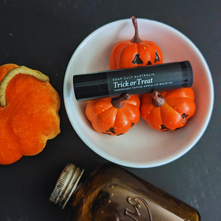 trick or treat lip balm in white bowl with orange jackolanterns near a plush velvet pumpkin and antique amber bottle on mottled black surface